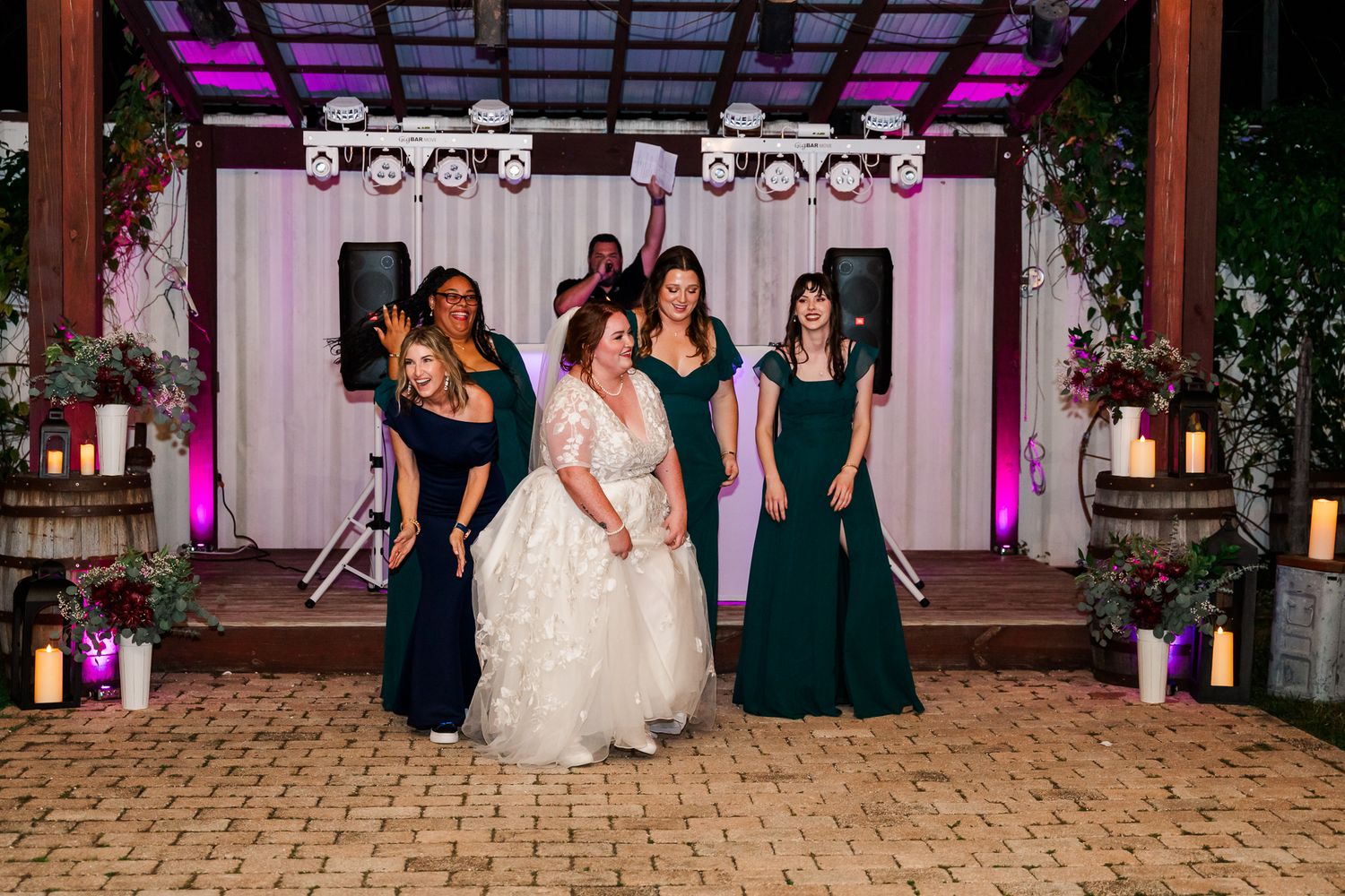 Wedding party members dance and celebrate during an evening reception at a venue with pink and purple lighting.