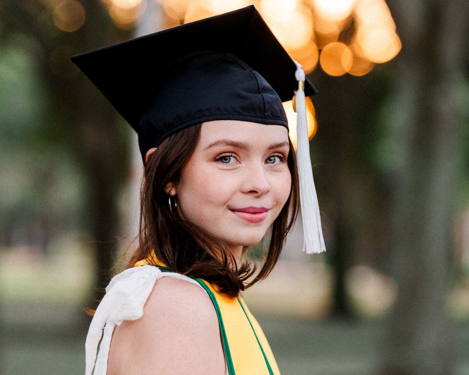 Graduate with dark hair in graduation cap and tassel poses for portrait series outdoors at sunset.