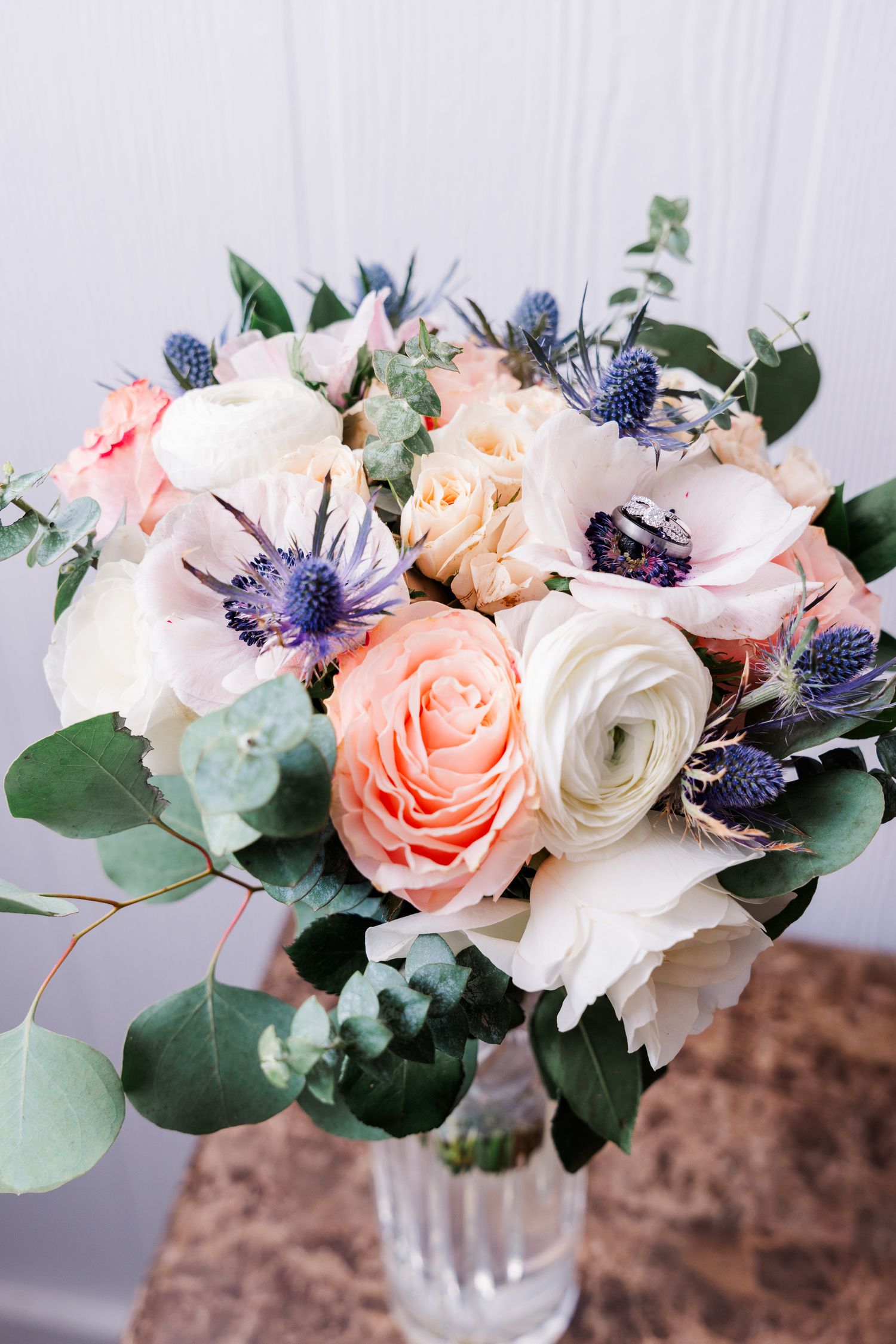 Elegant wedding bouquet with peach roses, white flowers, and blue thistle against white background.