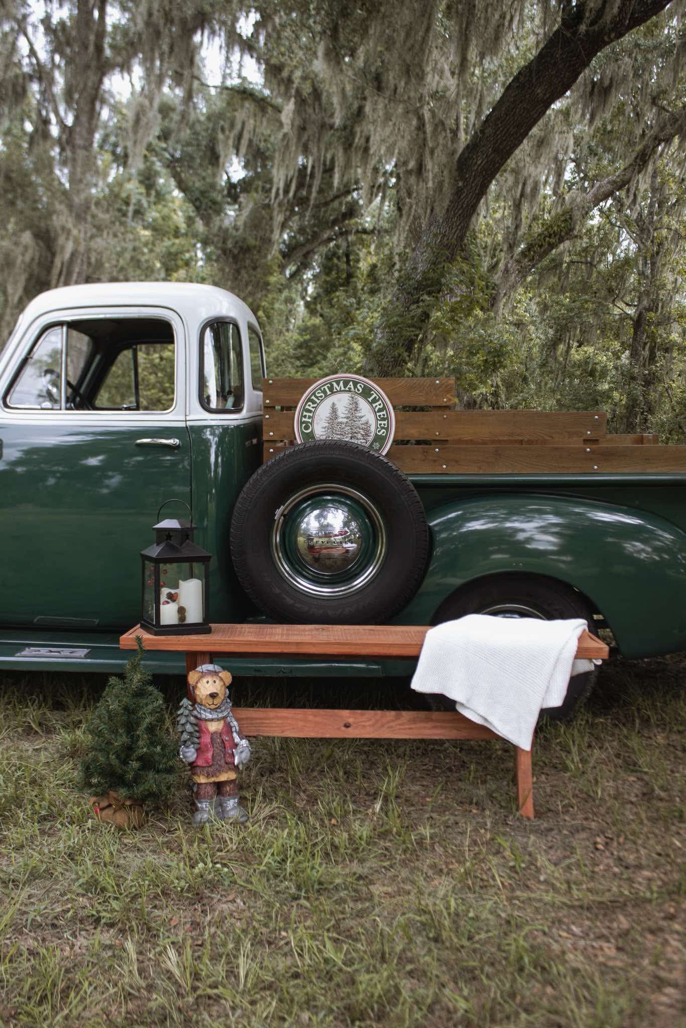 Vintage green pickup truck with wooden bench, Christmas bear figurine, lantern, and white blanket under Spanish moss trees.