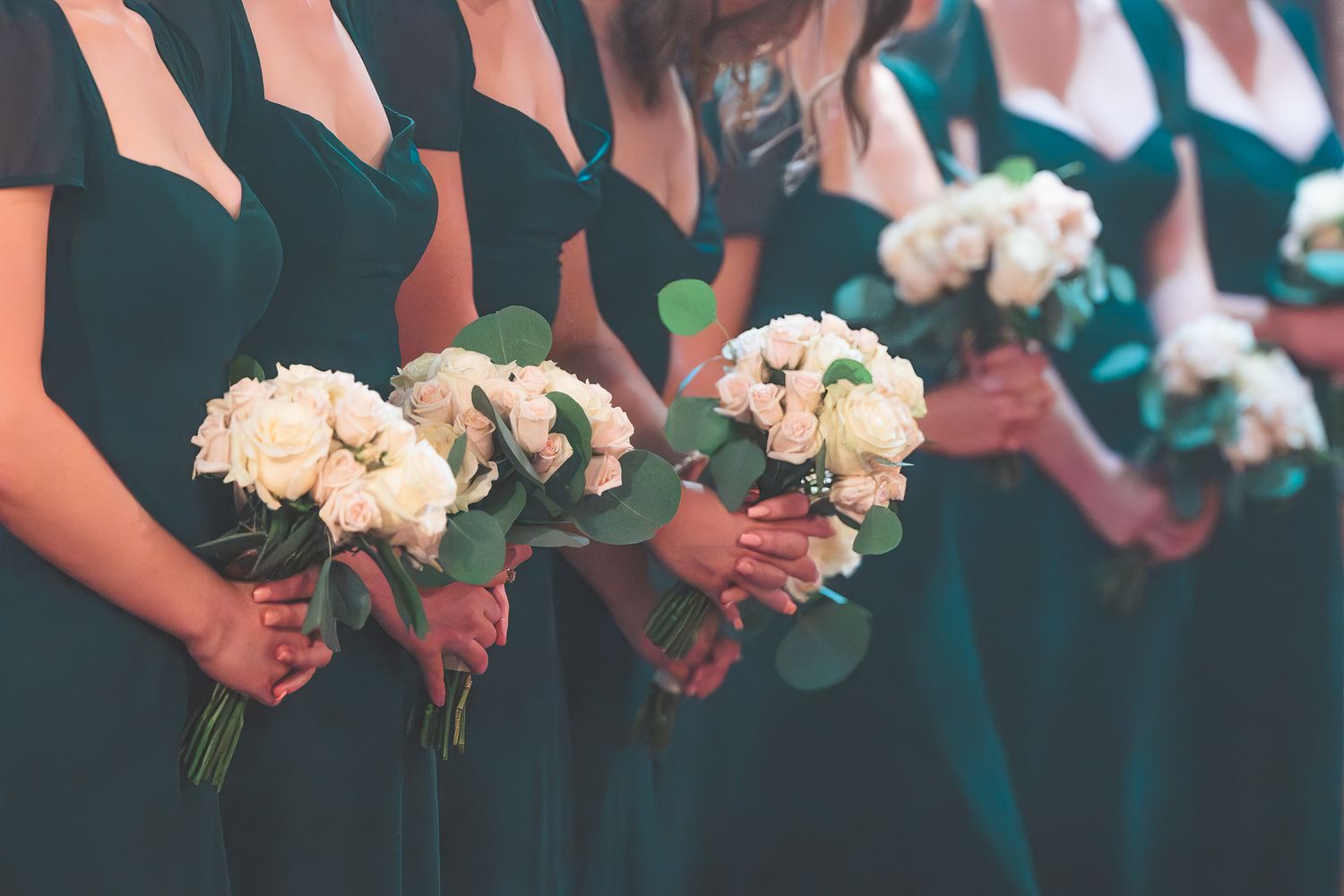 Row of wedding party members holding white rose bouquets against a dark background.