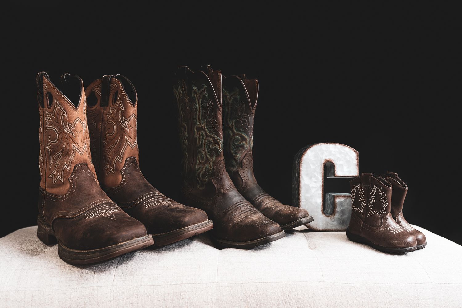 Family sized collection of brown leather cowboy boots arranged in descending size order on white background with letter C decor.