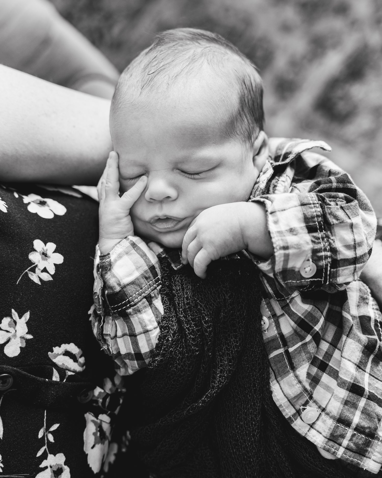 Sleeping baby in plaid shirt rests peacefully against floral print fabric in black and white portrait.