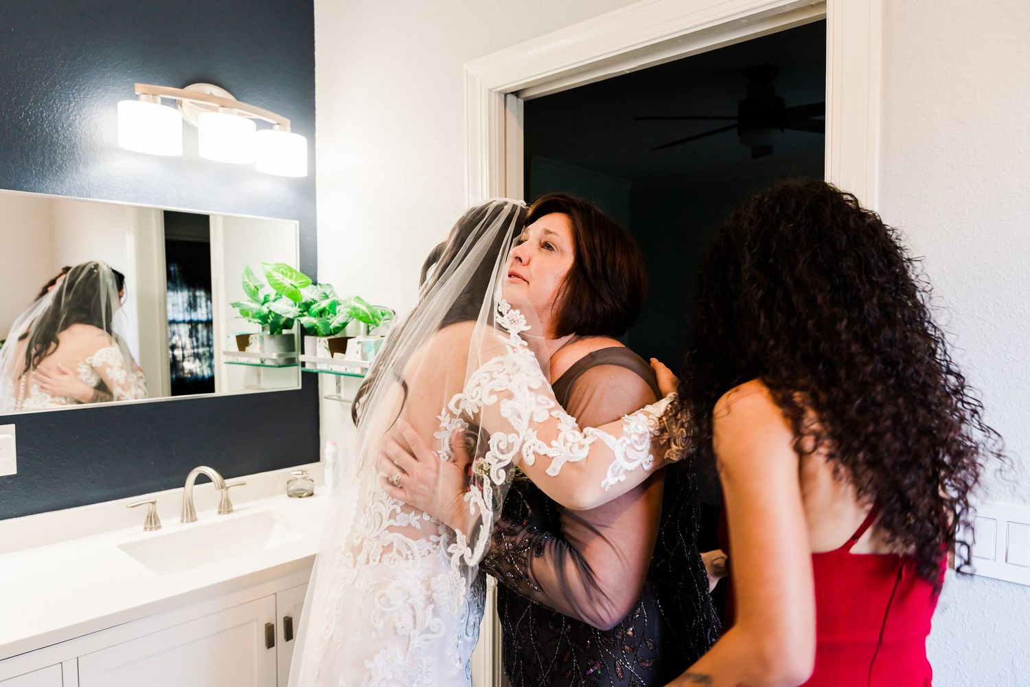 Friends help someone try on a white lace wedding dress in a bathroom with a white vanity and mirror.