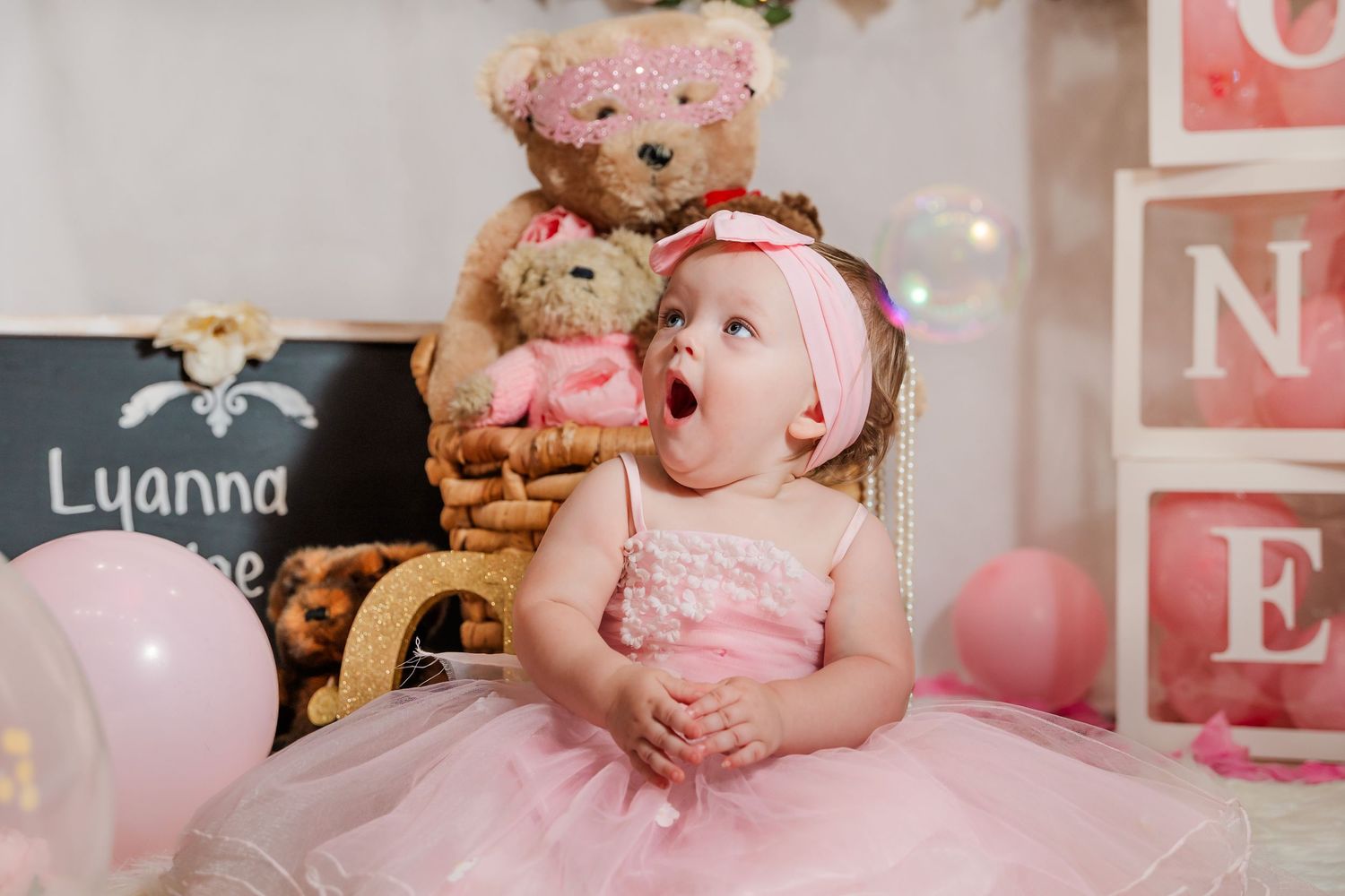 A one-year-old in a pink tulle dress poses for a milestone photo with teddy bears and alphabet blocks in the background.