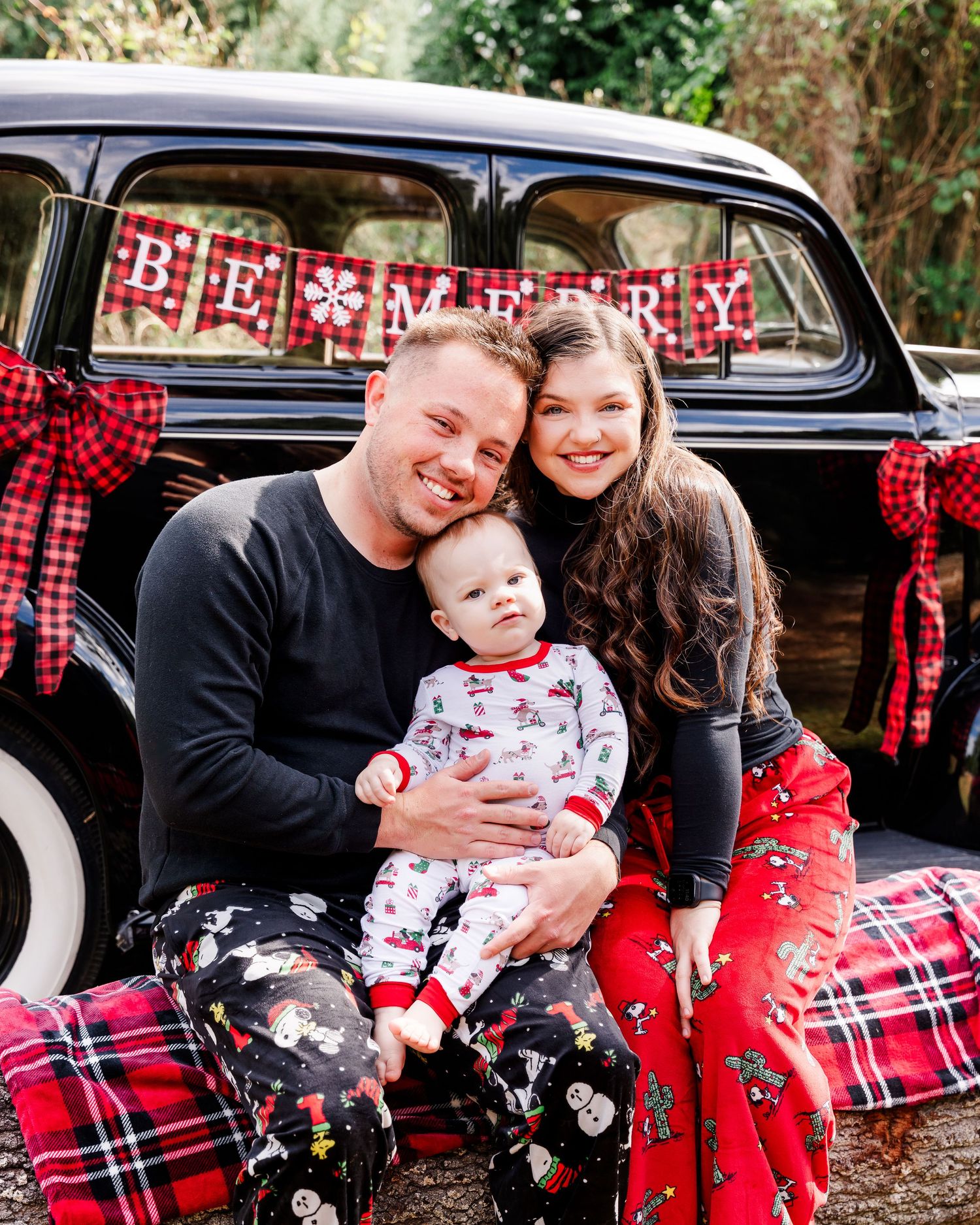 Family of three in Christmas pajamas posing with a vintage black car decorated with red plaid bows and a Be Merry banner.