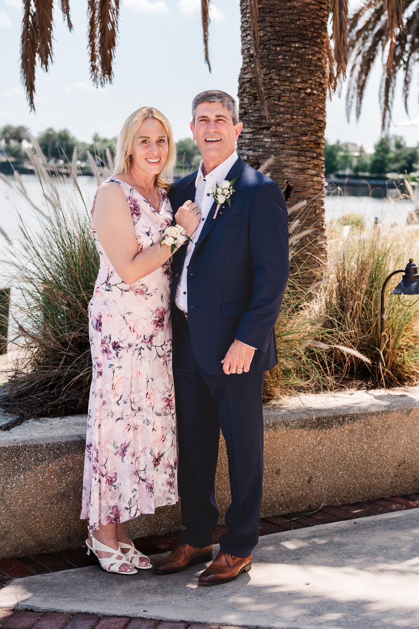 A couple poses together near a palm tree by the water, wearing formal attire including a pink floral dress and navy suit.