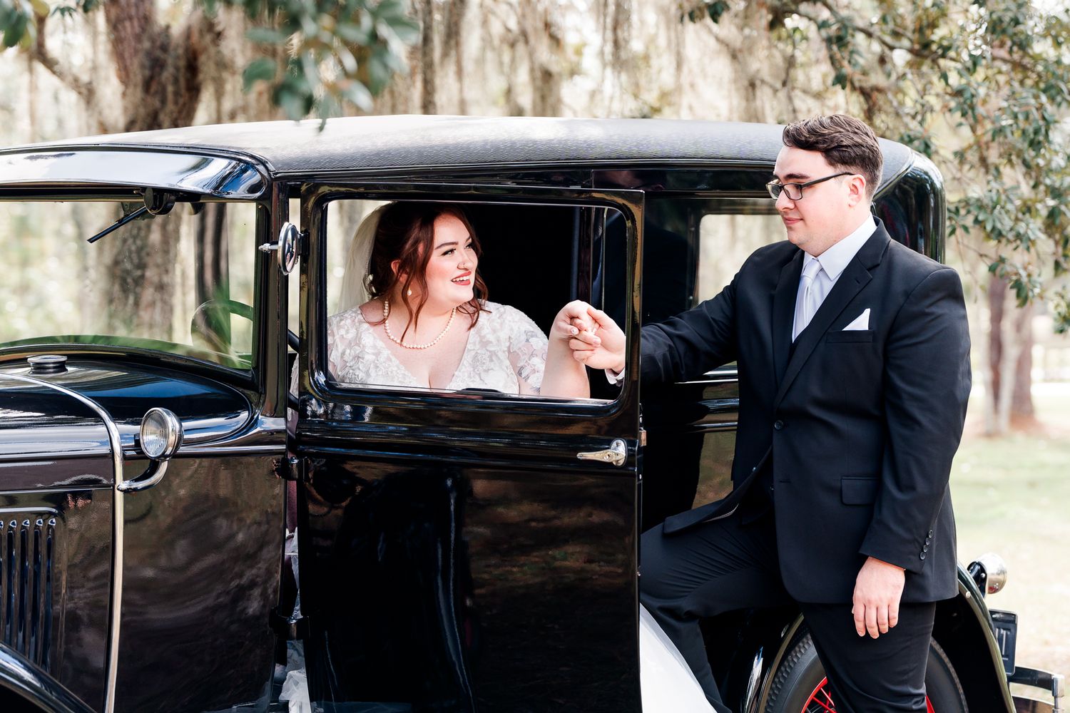 A vintage black car parked outdoors near trees serves as a backdrop for a romantic moment.