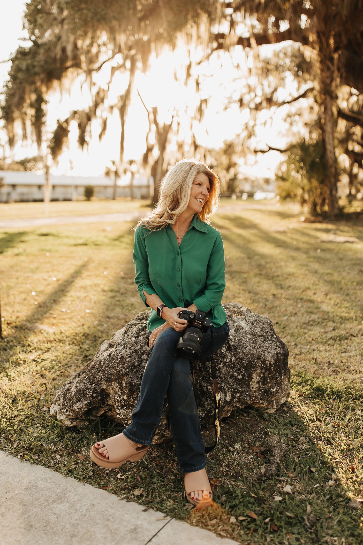 Someone in a green button-down shirt and jeans sits on a large rock at sunset with Spanish moss hanging from trees above.