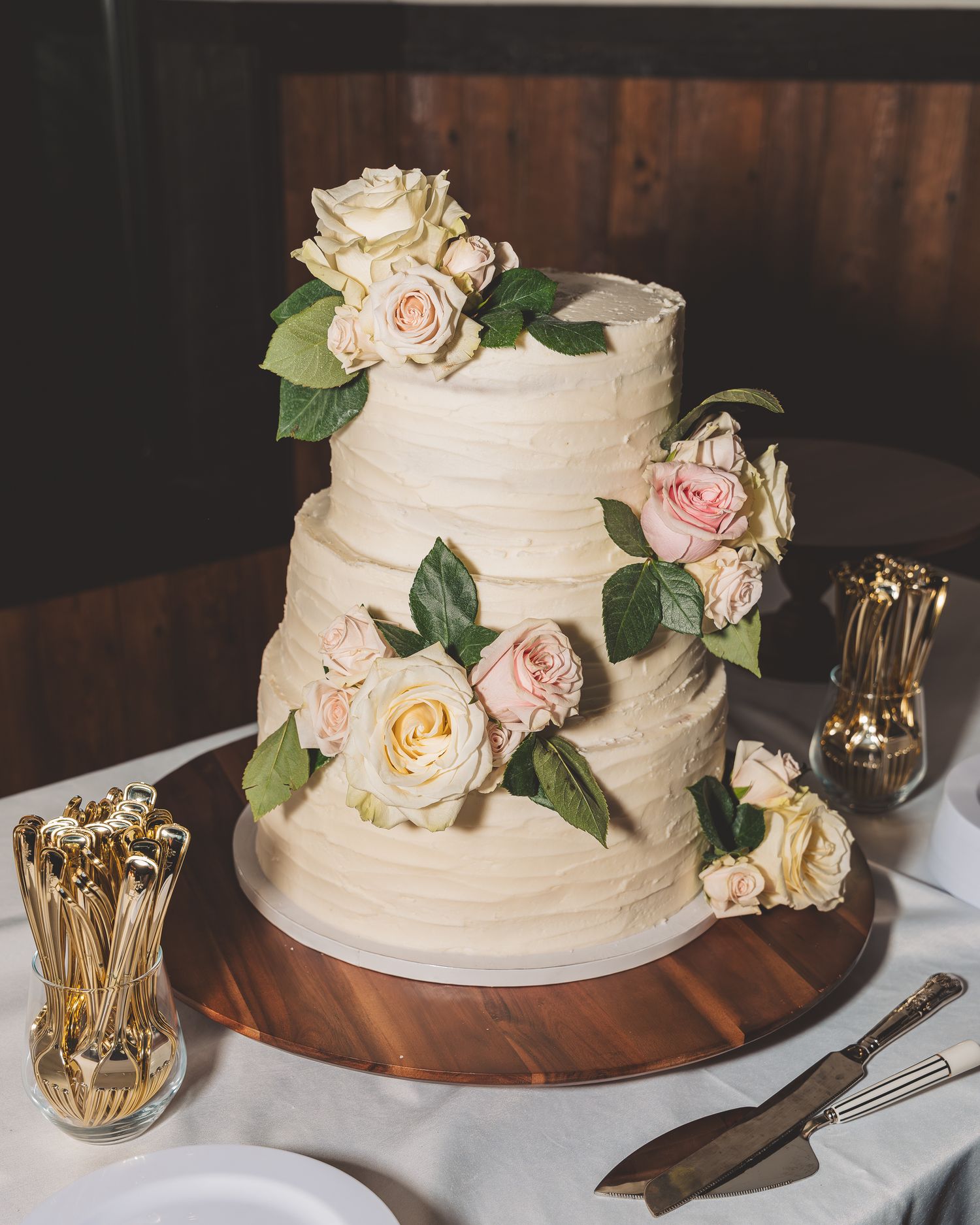 Three-tiered white wedding cake decorated with fresh pink and cream roses on a wooden stand.