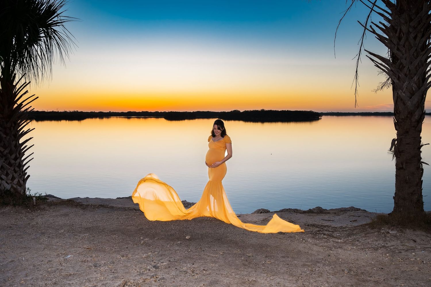 A silhouette stands on a beach at sunset wearing a flowing yellow dress as palm trees frame the peaceful water view.