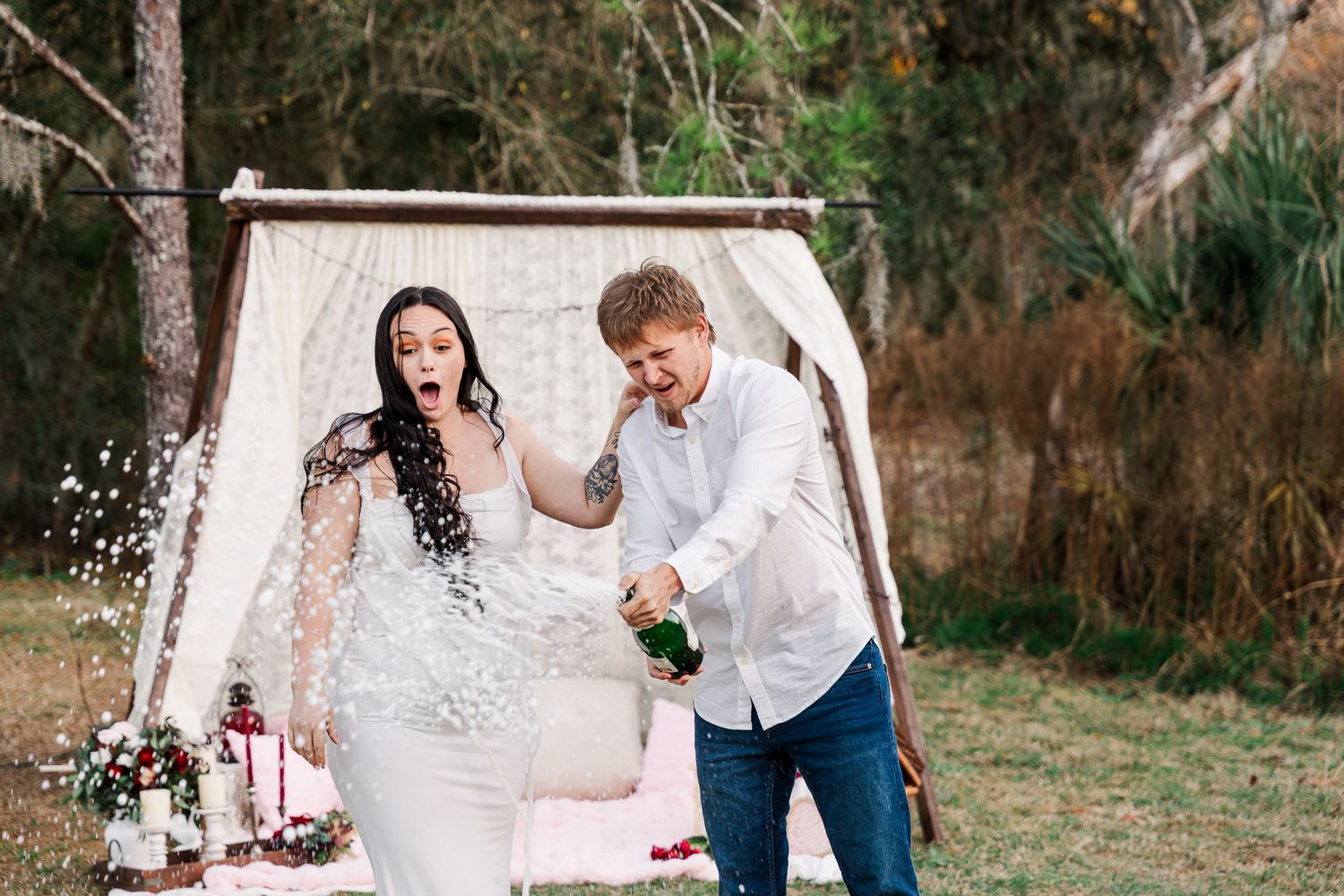 A couple in wedding attire pops champagne and celebrates in an outdoor garden setting.