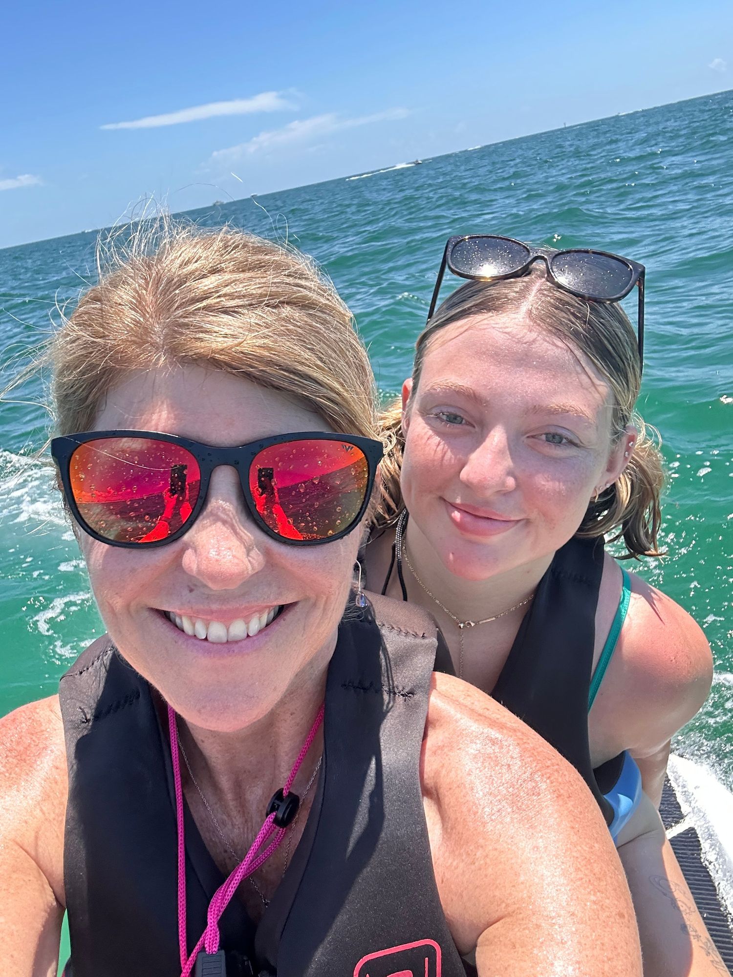 Two people wearing sunglasses take a selfie while jet skiing on turquoise ocean waters.