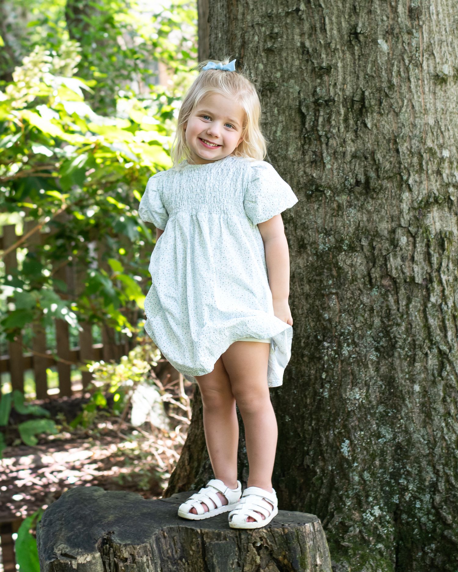 Smiling toddler girl in white floral dress and sandals standing on a tree stump outdoors.