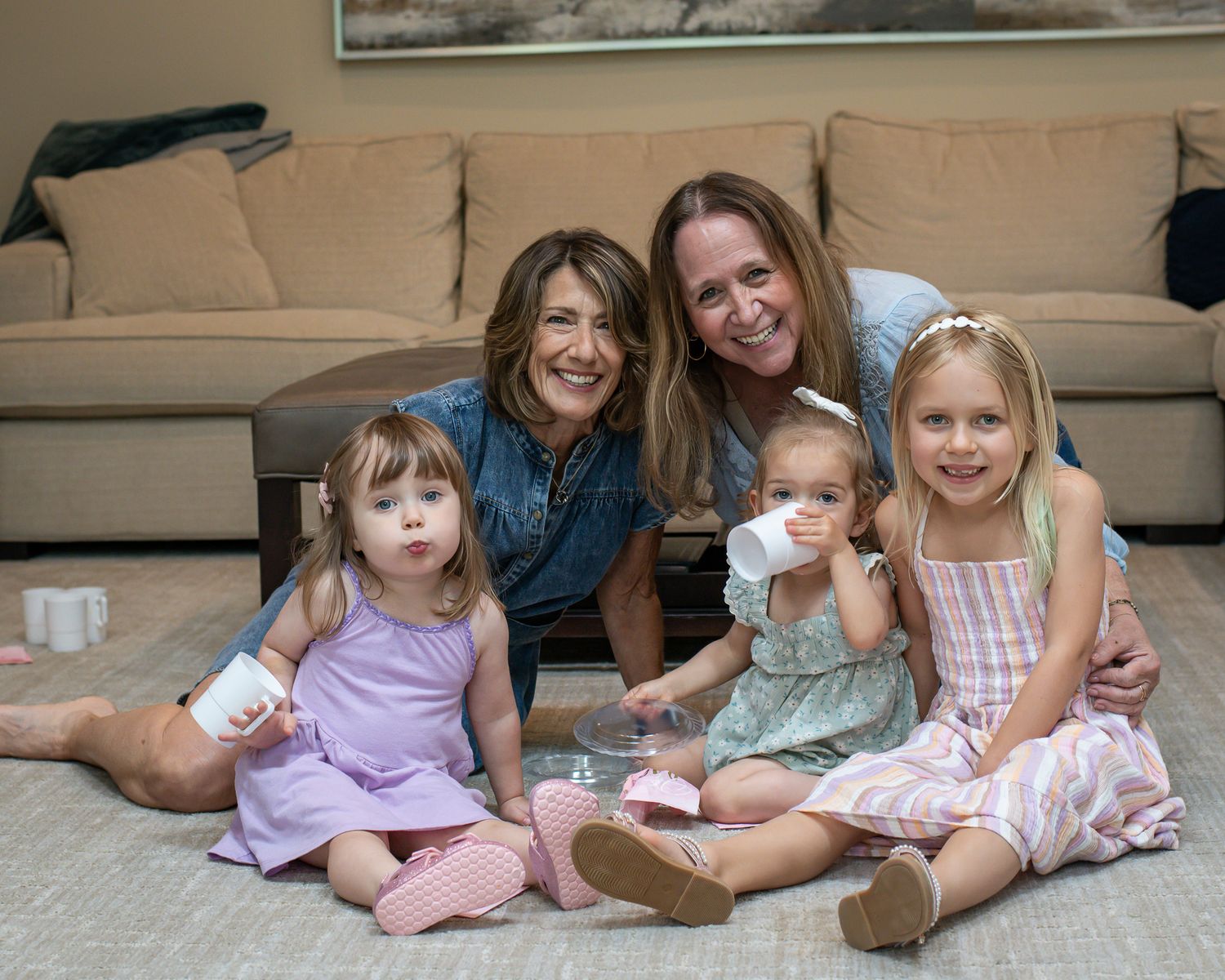 Two women and three young girls smile together on a living room floor in front of a beige couch.