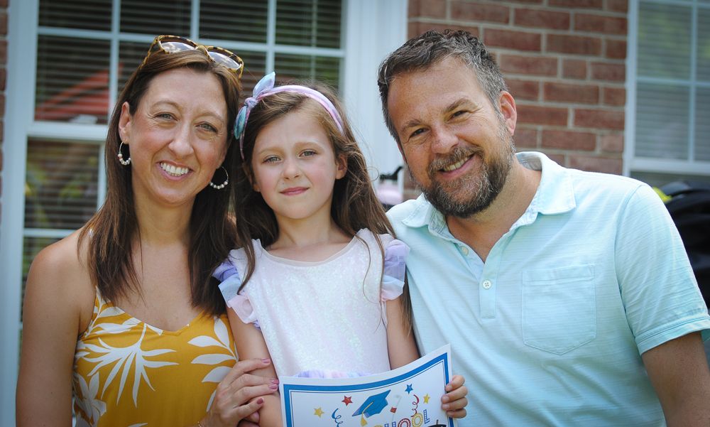 Family poses together outdoors smiling with a brick wall and windows in the background.