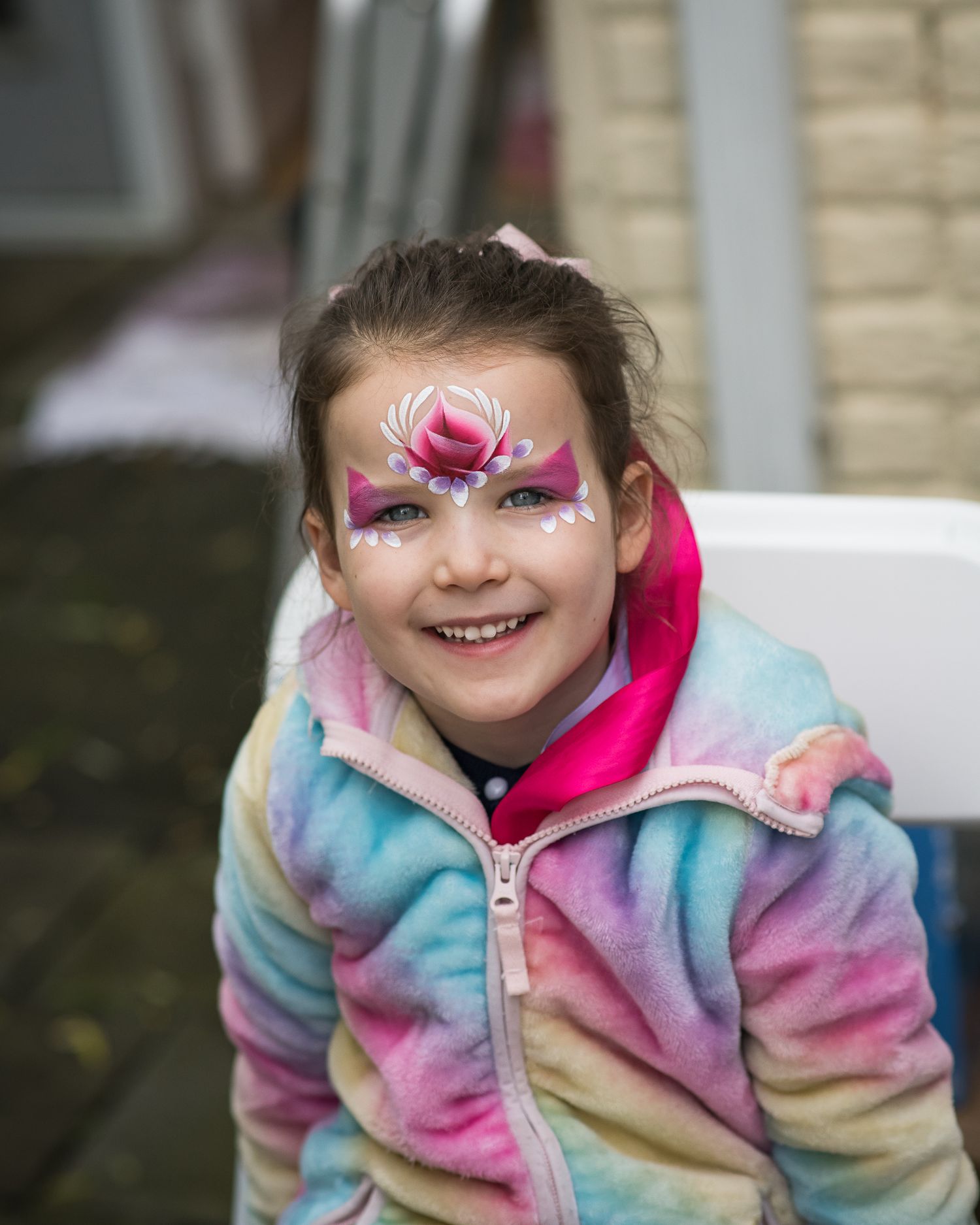 Smiling girl with pink butterfly face paint wearing a colorful tie-dye fleece jacket outdoors.