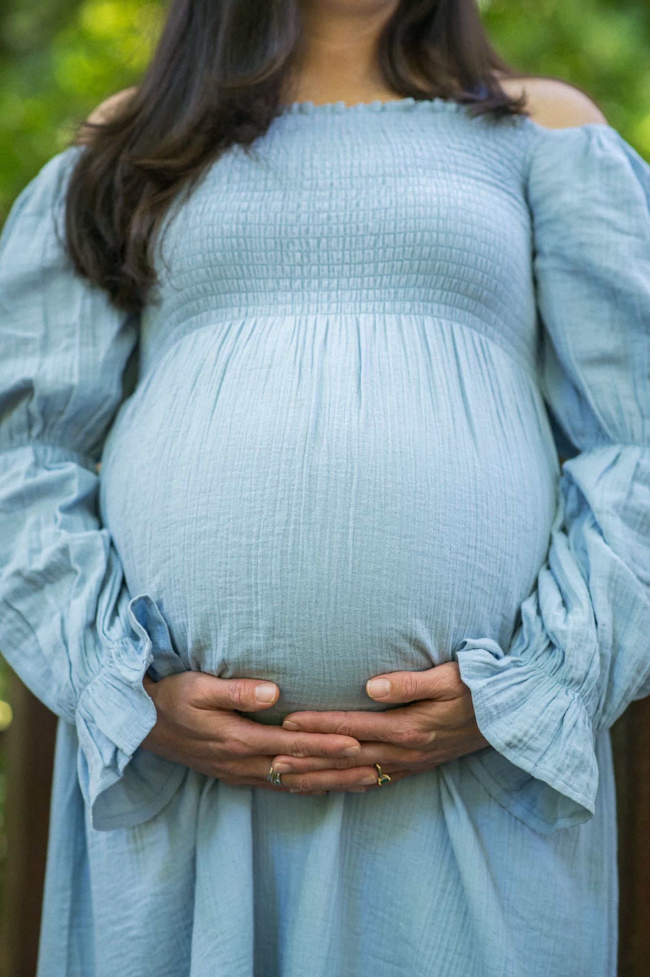 Pregnant woman in light blue smocked off-shoulder dress cradling her baby bump outdoors.