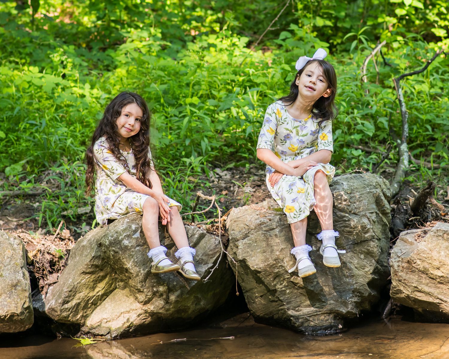 Two young girls in matching floral dresses sitting on rocks by a creek surrounded by lush green trees.