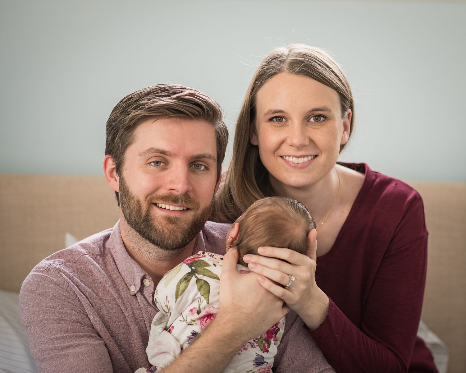 A couple in burgundy and pink sweaters holding a newborn baby in a floral outfit for a family portrait.