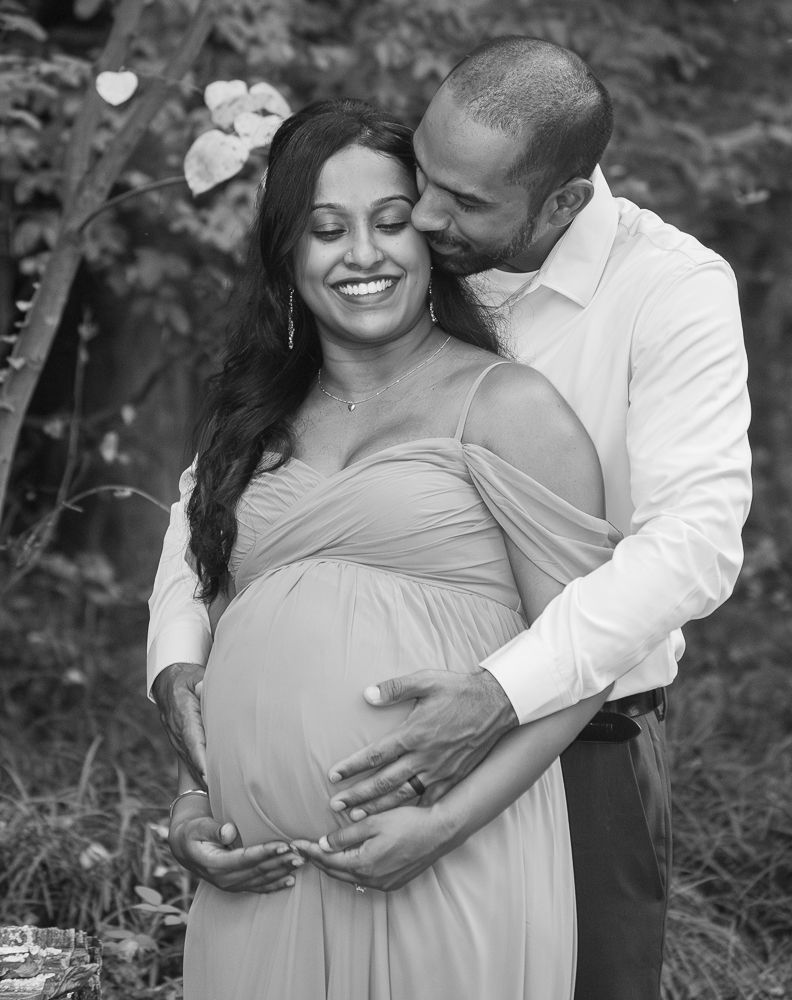 Black and white maternity photos showing a couple embracing outdoors in a romantic woodland setting.