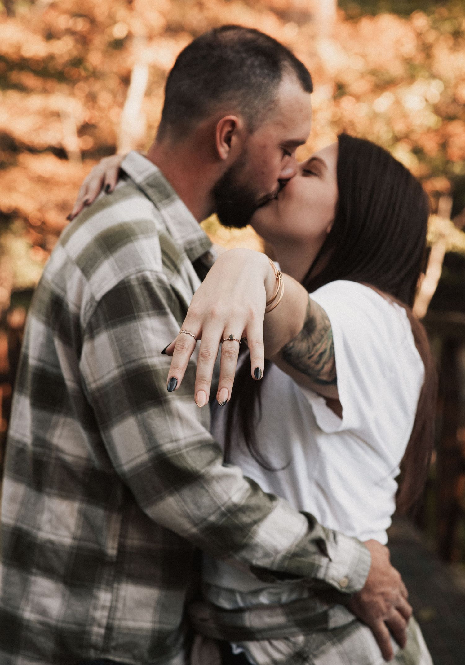 Couple sharing a romantic kiss outdoors in autumn, woman displaying engagement ring on hand around man's neck.