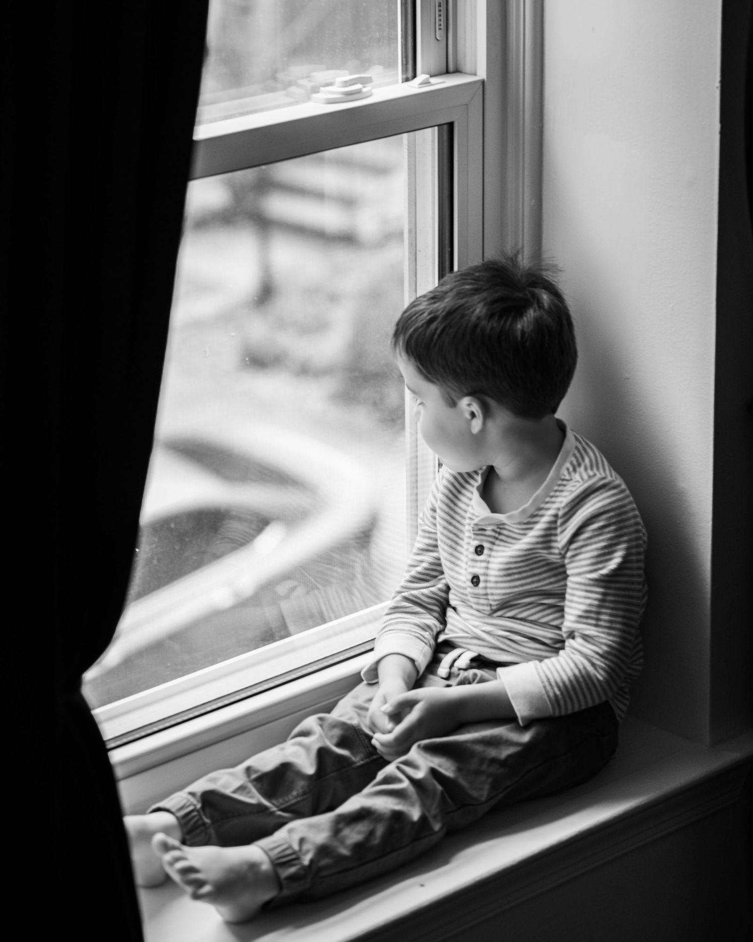 Black and white photo of a young boy sitting on a windowsill, gazing thoughtfully outside through a large window.