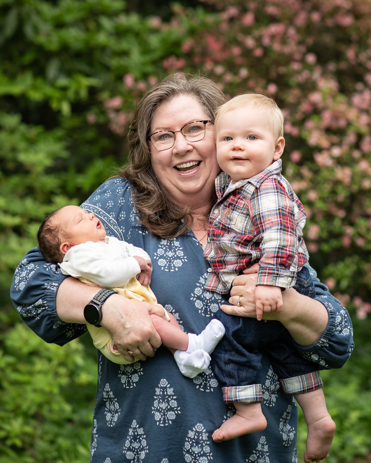 Smiling woman holding a newborn baby and a toddler boy outdoors in a lush green garden setting.