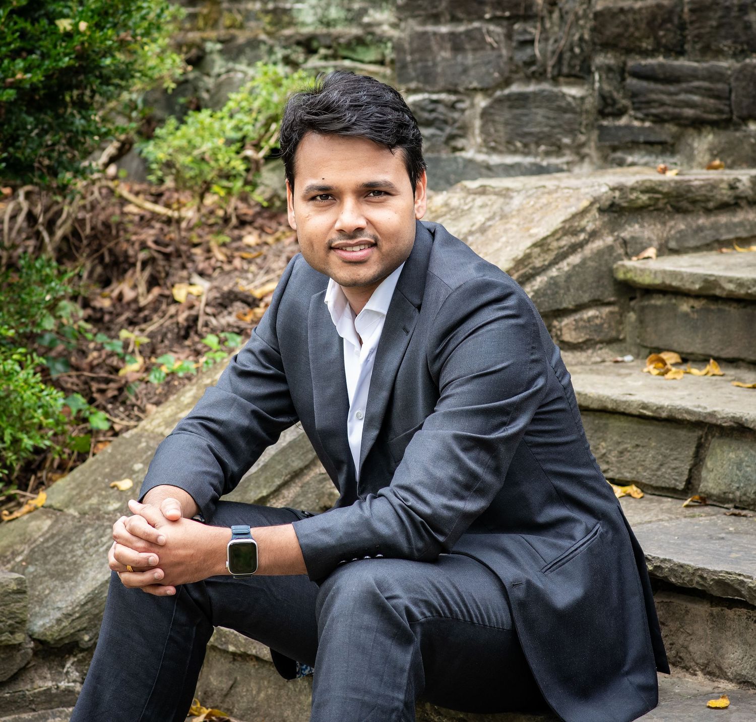 A person in a dark suit sits casually on stone steps outdoors surrounded by greenery and stone walls.