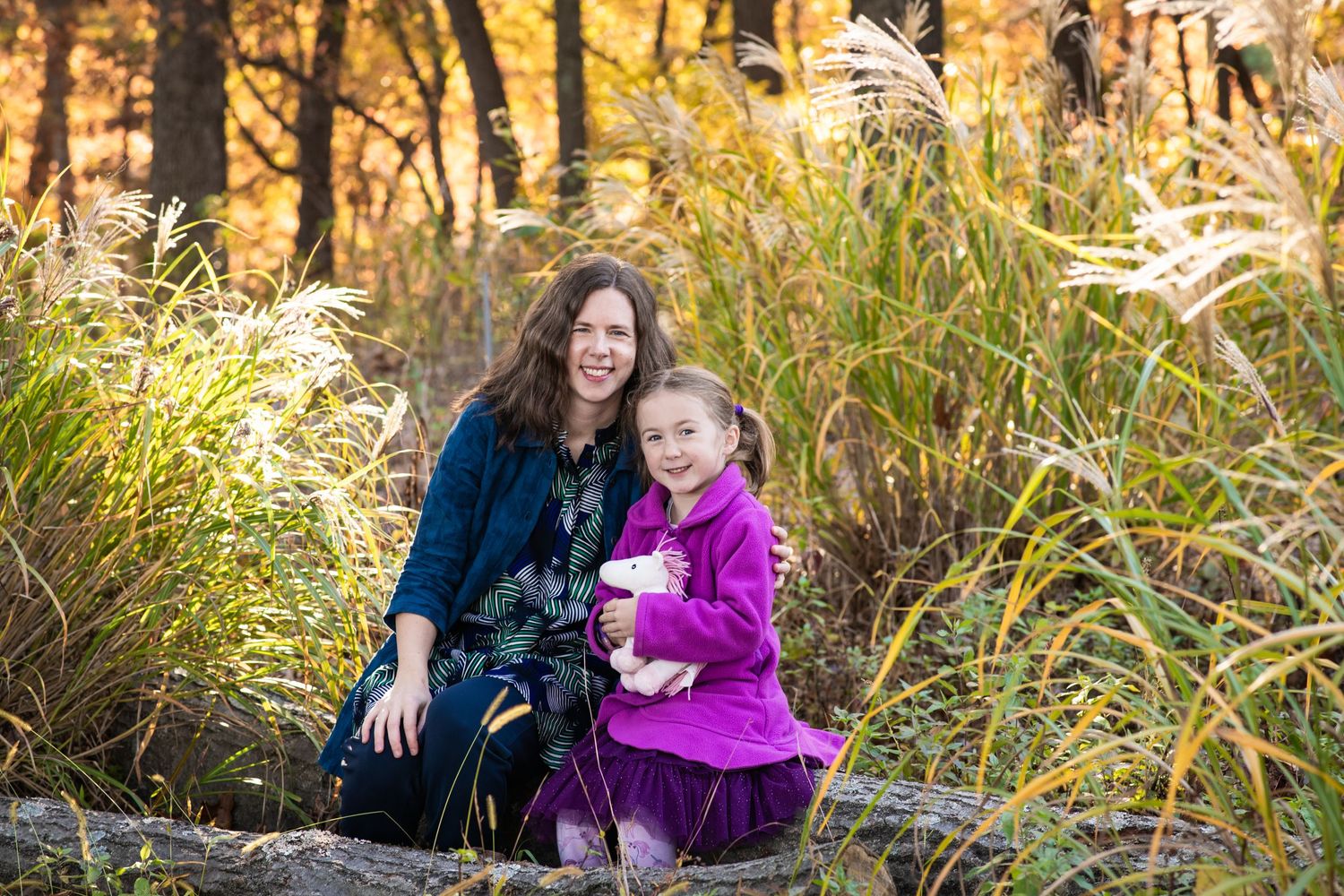Parent and child sit together in tall autumn grass during golden hour as sunlight filters through the trees.