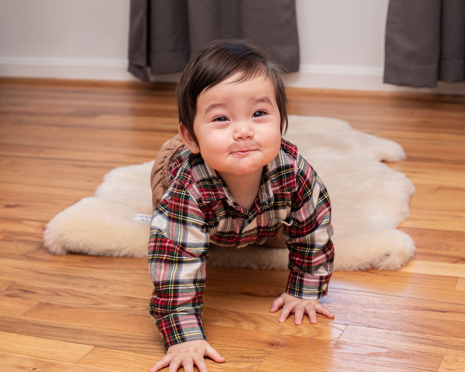 A child in a plaid shirt crawls playfully across a hardwood floor near a white fluffy rug.