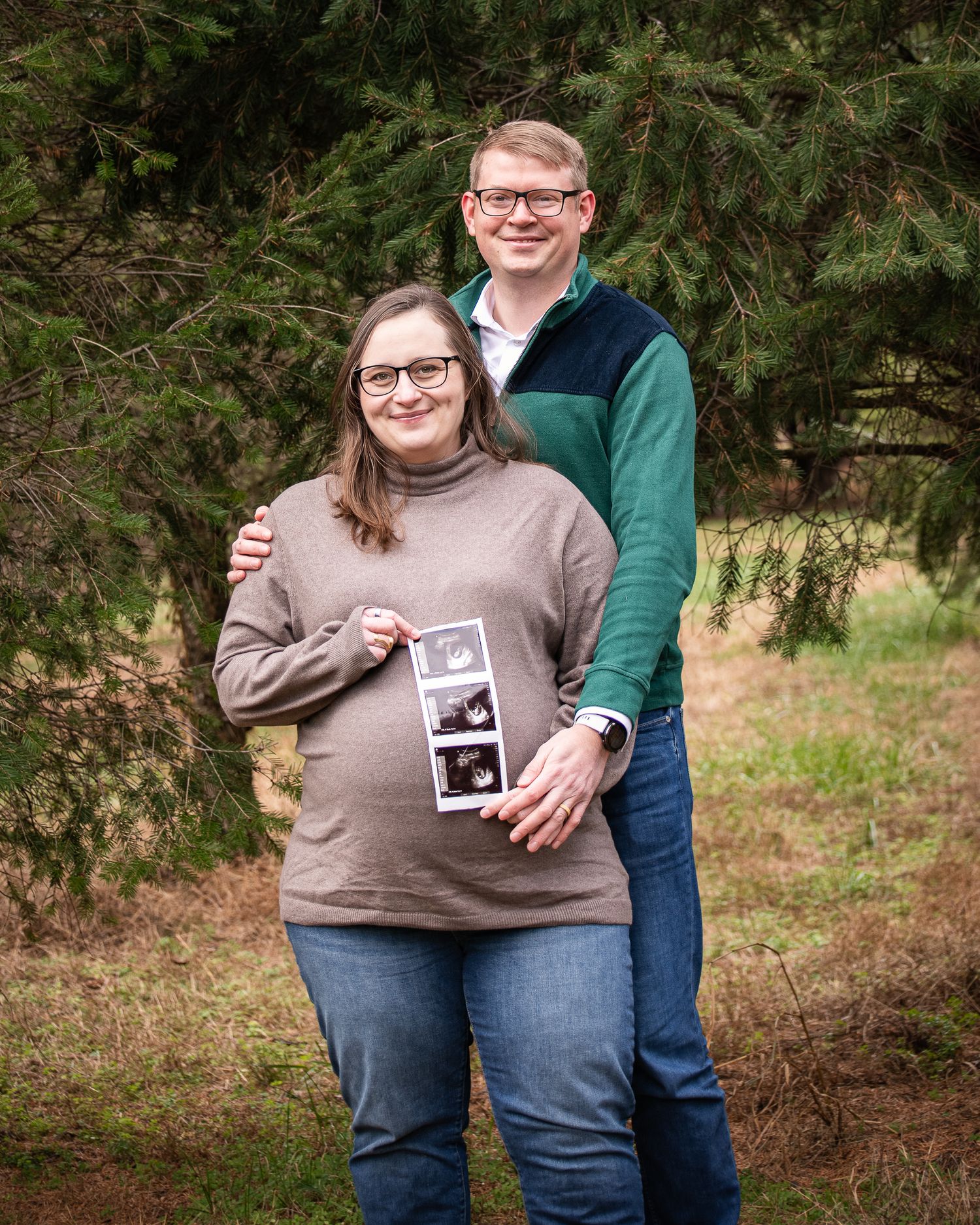 A couple in casual clothing stands outdoors near evergreen trees while holding a sonogram photo.