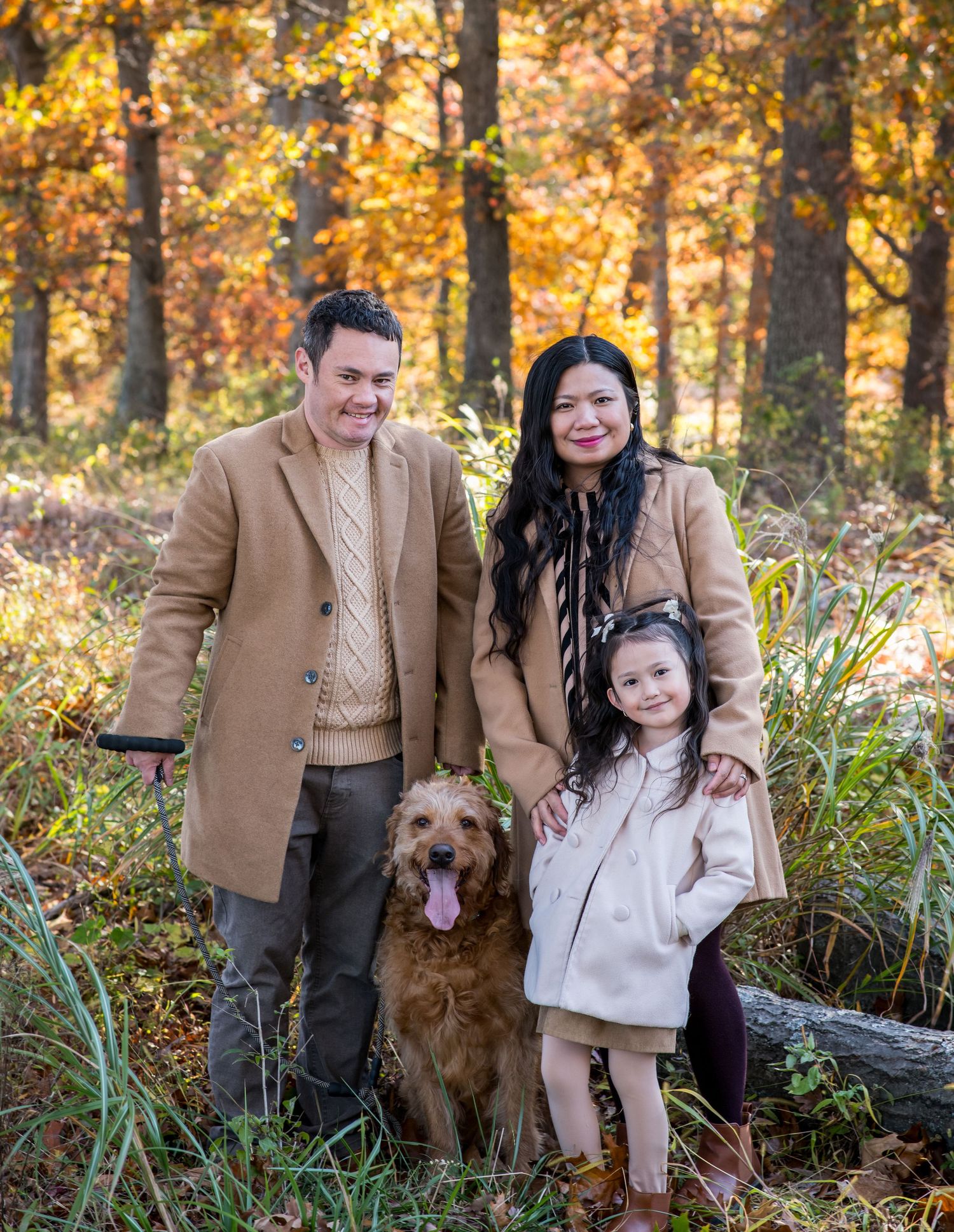 A family and their golden retriever pose together in a scenic autumn forest setting with golden foliage in the background.