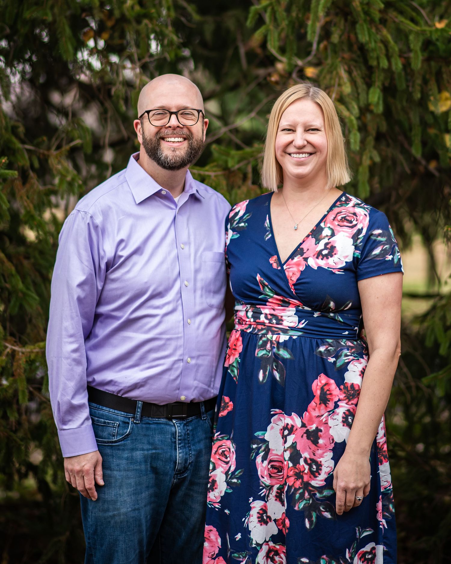 A couple poses together outdoors in coordinating purple shirt and floral dress against a natural backdrop.