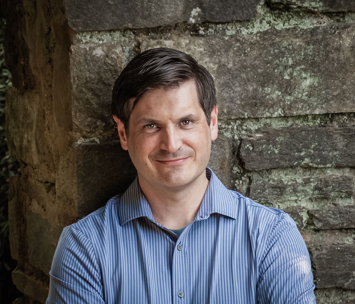 Professional headshots showing a person in a blue striped dress shirt against a textured stone wall background.