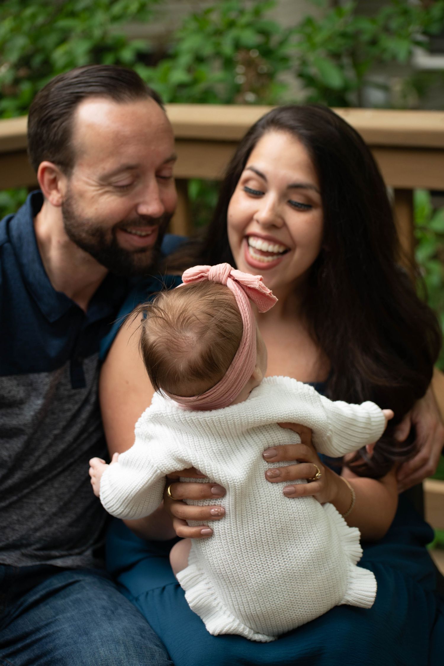 Happy couple smiling and laughing while holding their newborn baby in a white sweater outdoors.