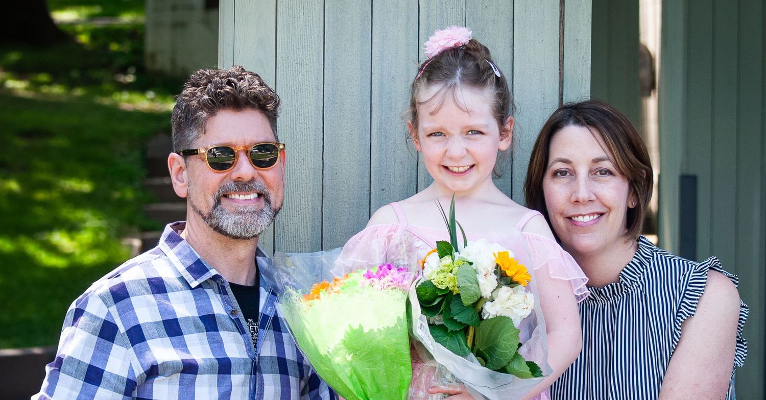 A young girl in a pink dress holds a bouquet of flowers, smiling with two adults outdoors.