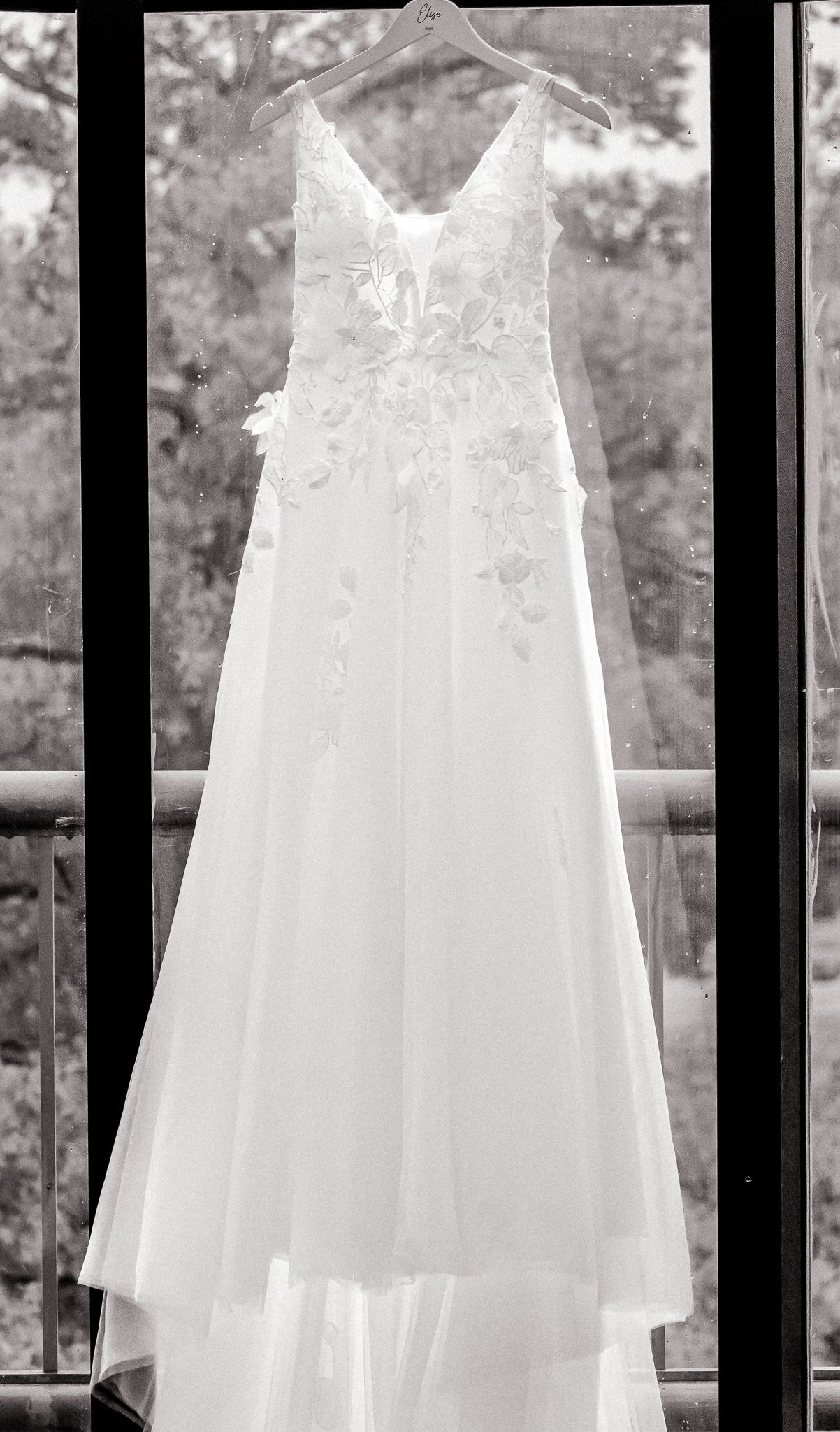 Black and white photo of an elegant lace wedding gown hanging on a hanger in front of a window.
