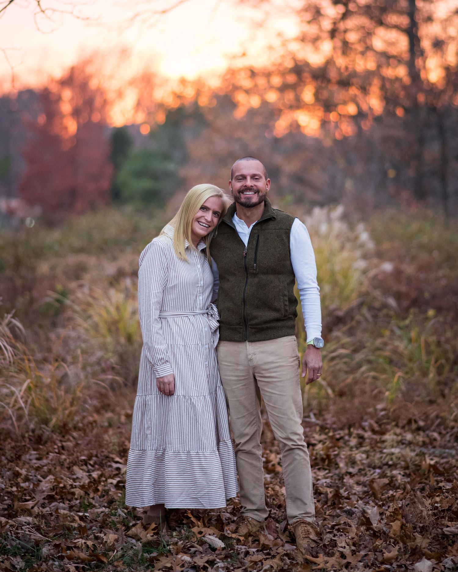 A couple walks together through fallen autumn leaves at sunset in a wooded area.