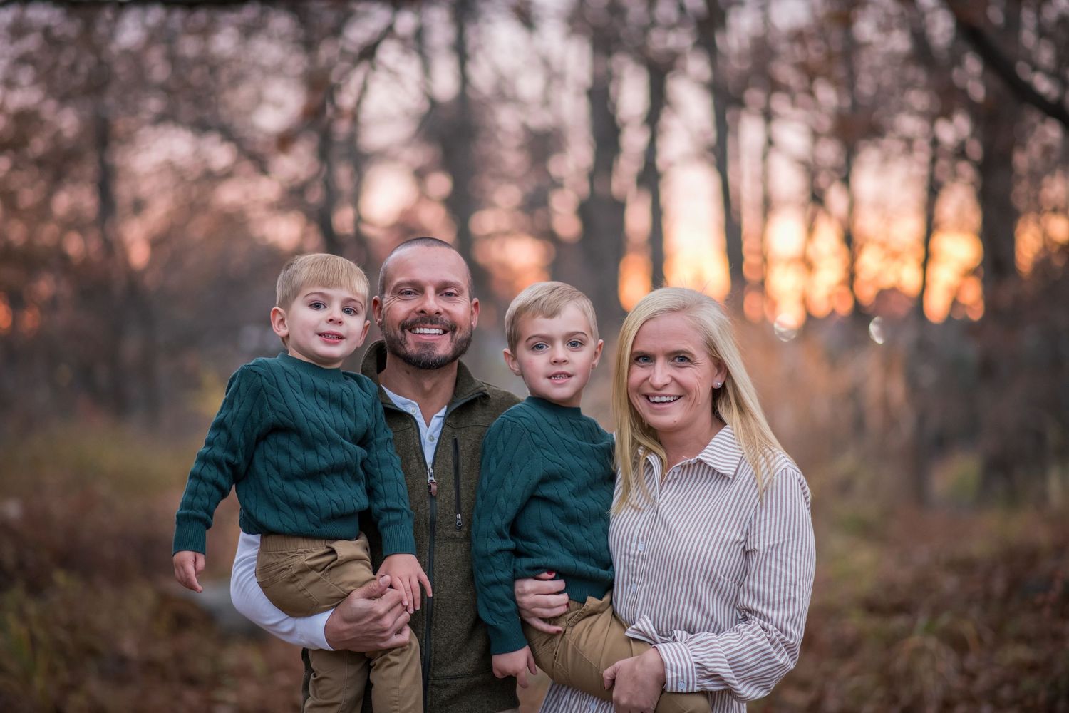 A family wearing coordinated green and white outfits poses together outdoors during sunset in a wooded setting.