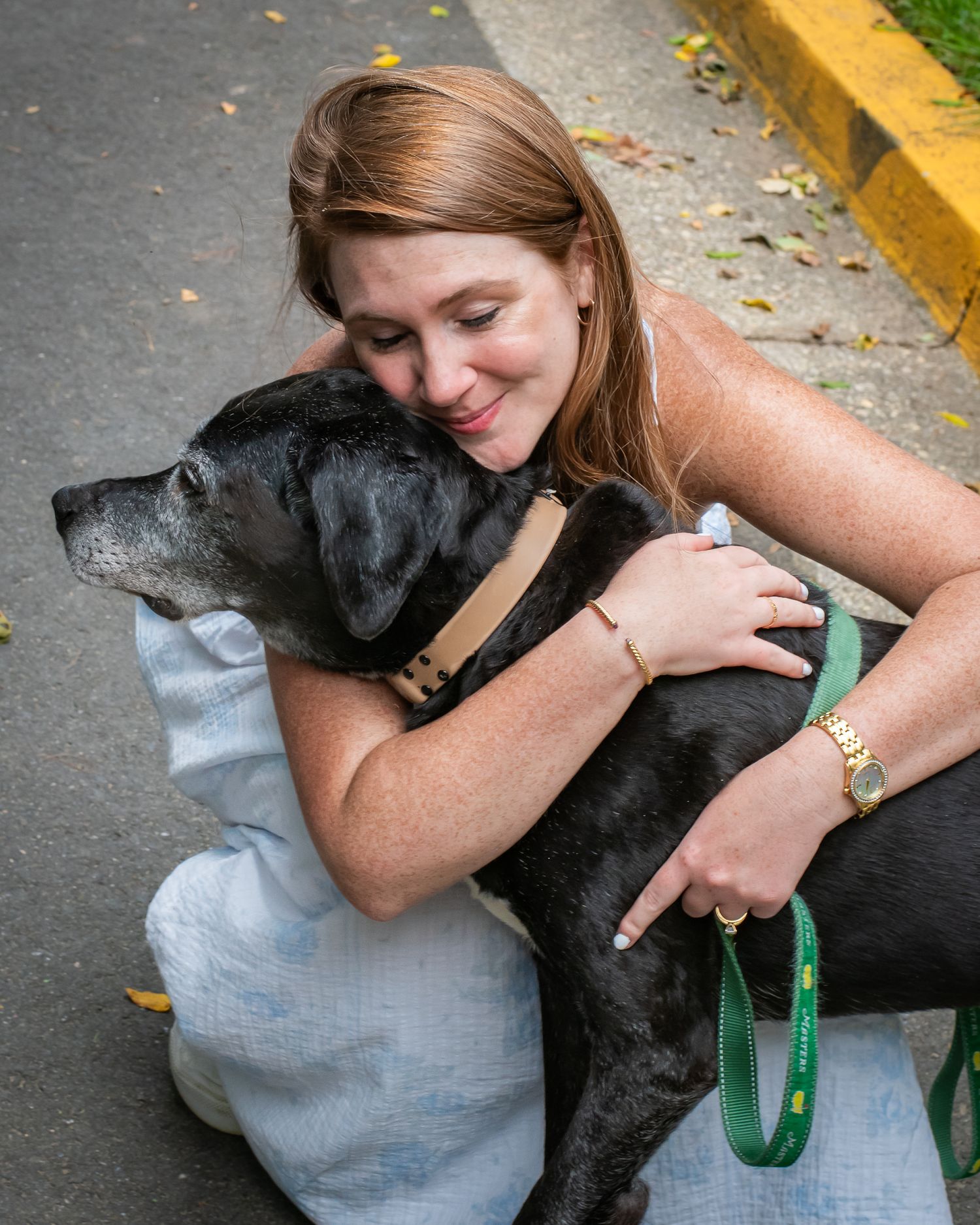 Woman with red hair hugging a black dog on a leash outdoors, smiling warmly while wearing a gold watch.