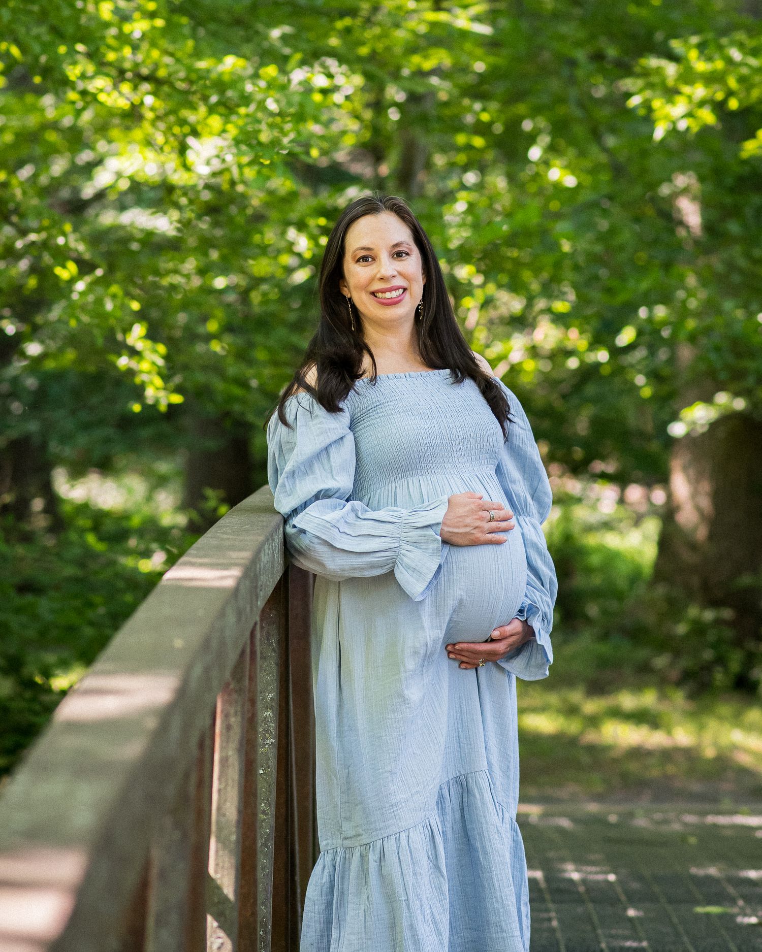 Smiling pregnant woman in light blue dress holding baby bump on wooden bridge surrounded by lush green trees.