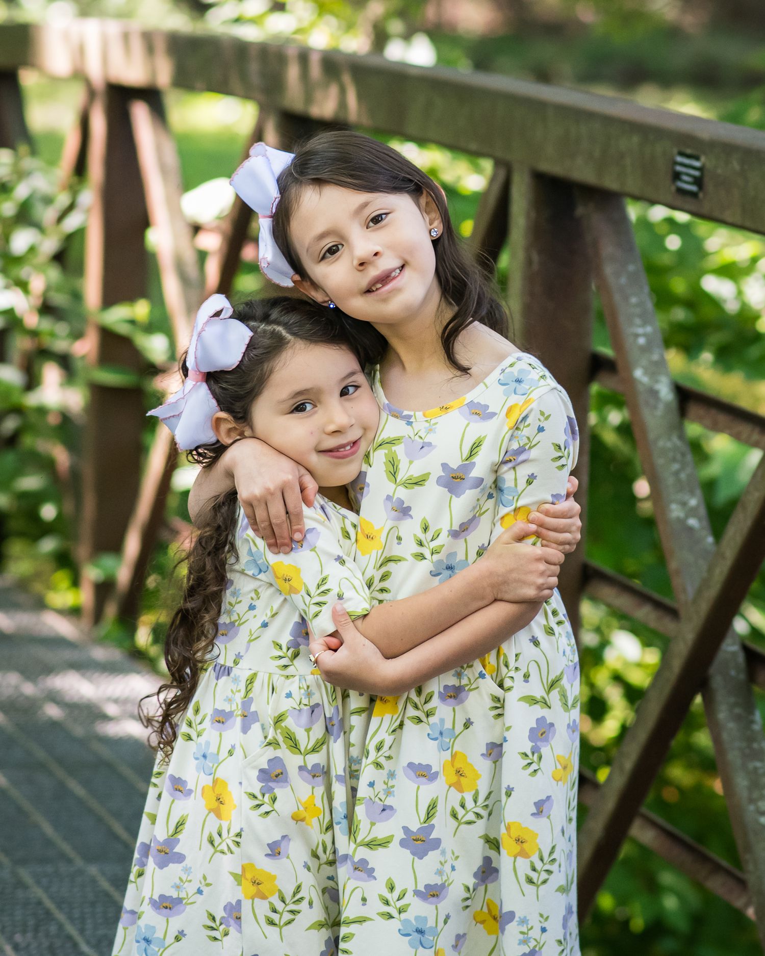 Two young girls in matching floral dresses embrace on a wooden bridge surrounded by lush greenery.