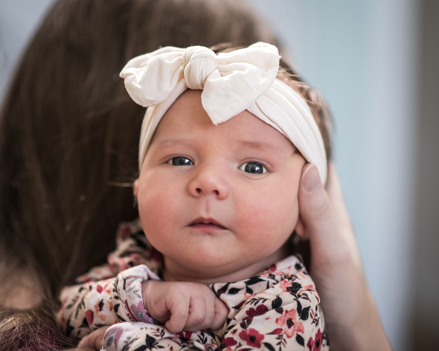 A baby wearing a white bow headband and floral outfit looks at the camera with wide-eyed curiosity.