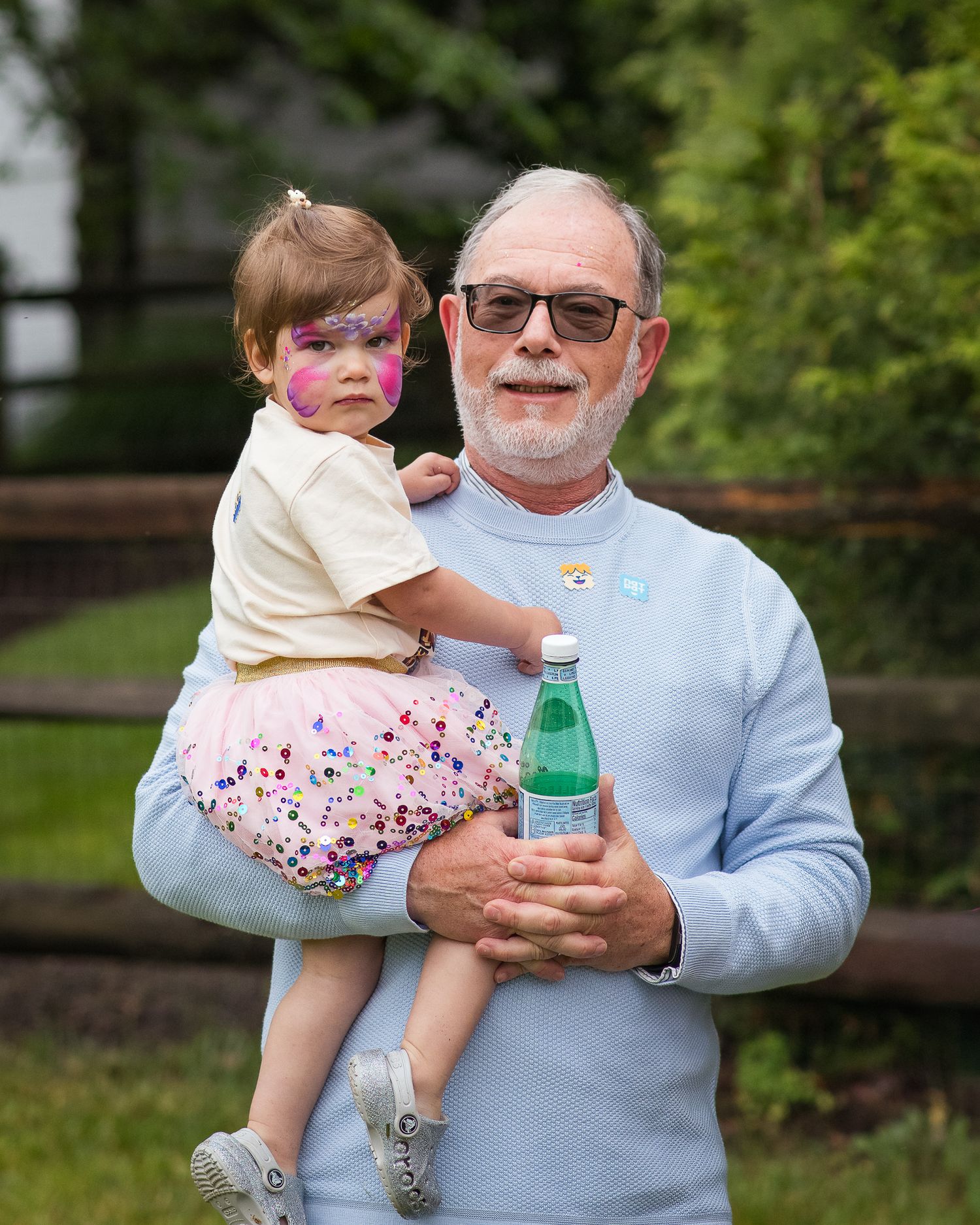 Elderly man with beard and glasses holds a toddler girl with face paint, outdoors near a wooden fence.