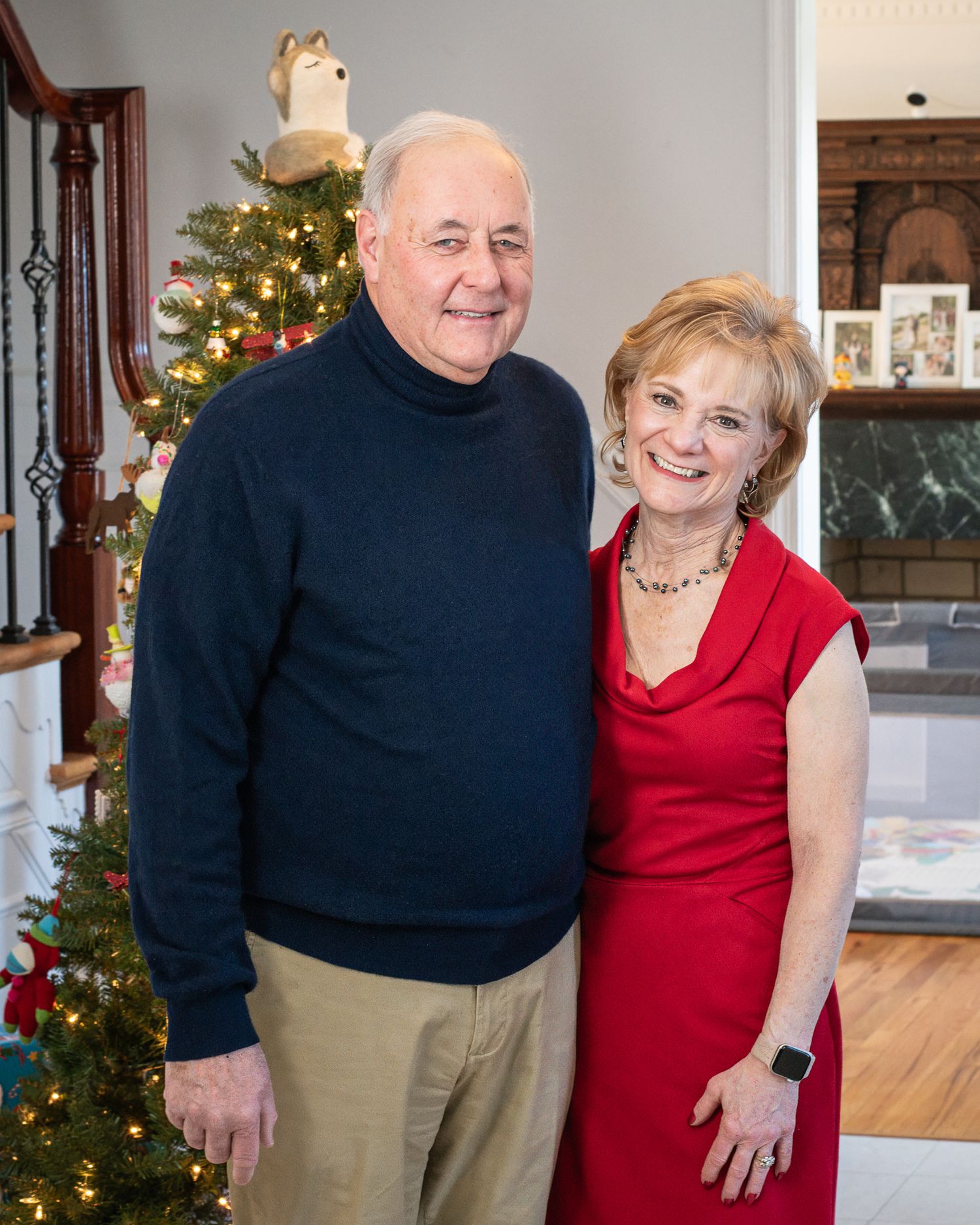 A couple poses in front of a decorated Christmas tree, wearing a navy sweater and red dress for a holiday photo.
