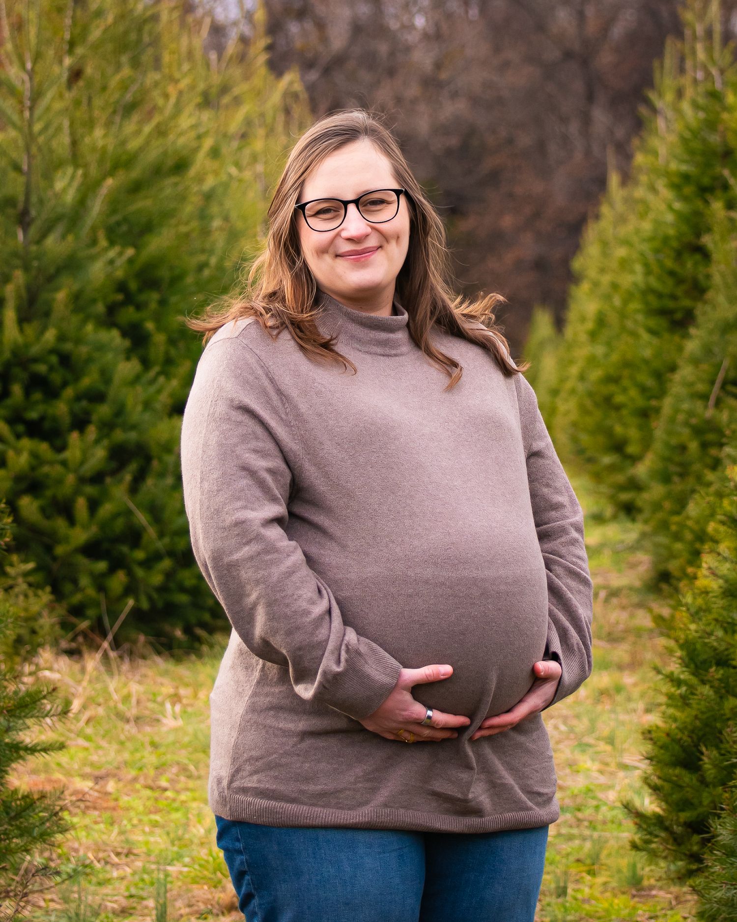 A person wearing a gray sweater stands among evergreen trees in an outdoor setting during winter.