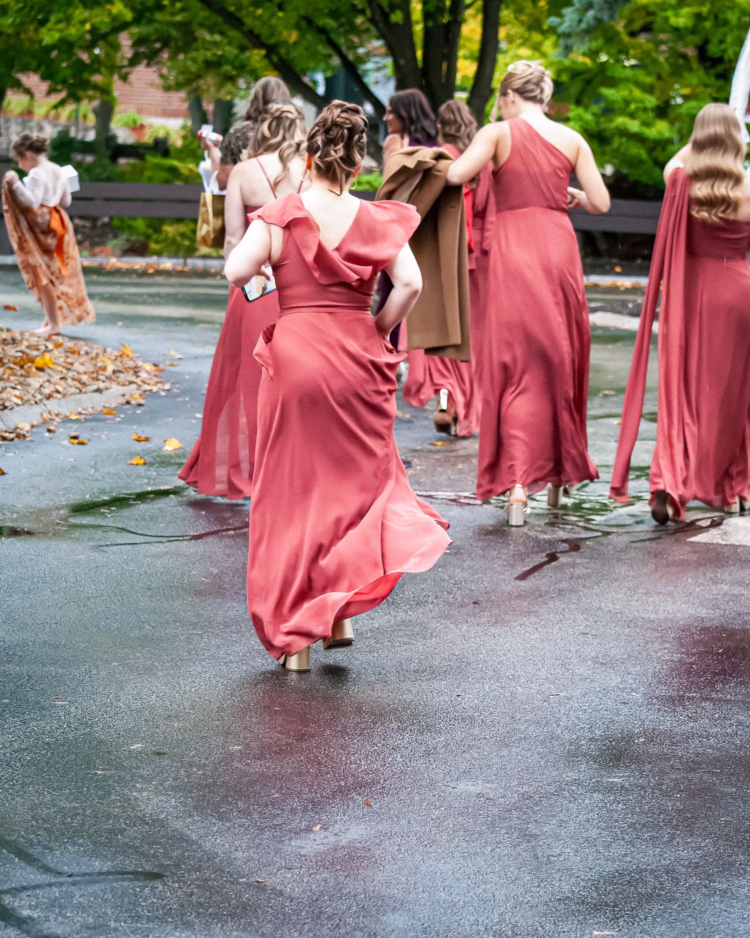 Bridesmaids in matching coral red gowns walking on wet pavement during an outdoor fall wedding ceremony.