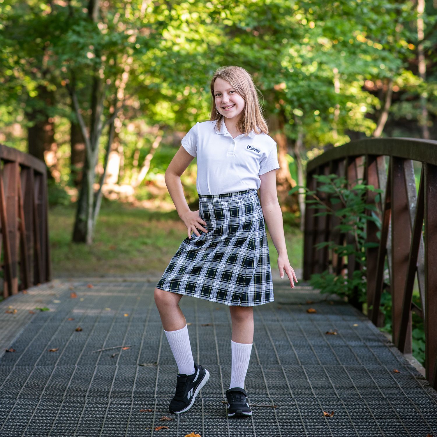 Young girl in school uniform with plaid skirt, white polo shirt, and knee socks poses on wooden bridge.