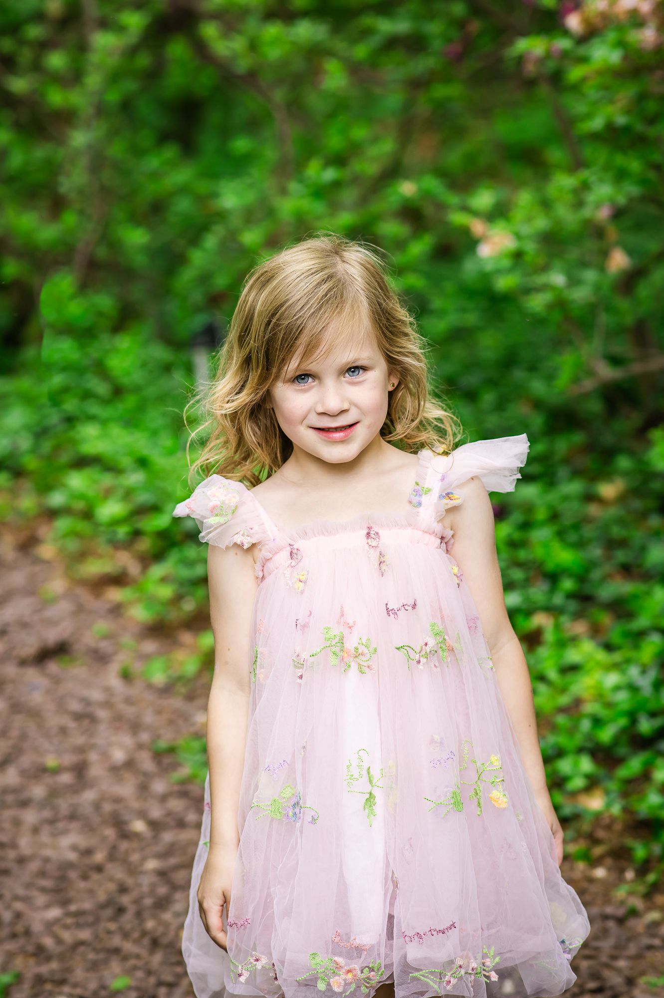 Young girl in pink floral tulle dress smiling outdoors among green trees and bushes.